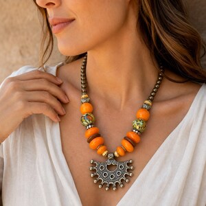 May include: A necklace featuring a central silver-toned metal pendant with a semi-circular, scalloped design. The chain consists of metallic grey beads, interspersed with bright orange spherical beads, brown disc beads, and green speckled beads.