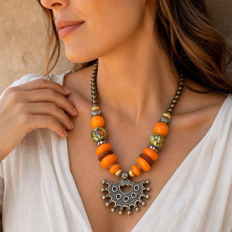 May include: A necklace featuring a central silver-toned metal pendant with a semi-circular, scalloped design. The chain consists of metallic grey beads, interspersed with bright orange spherical beads, brown disc beads, and green speckled beads.