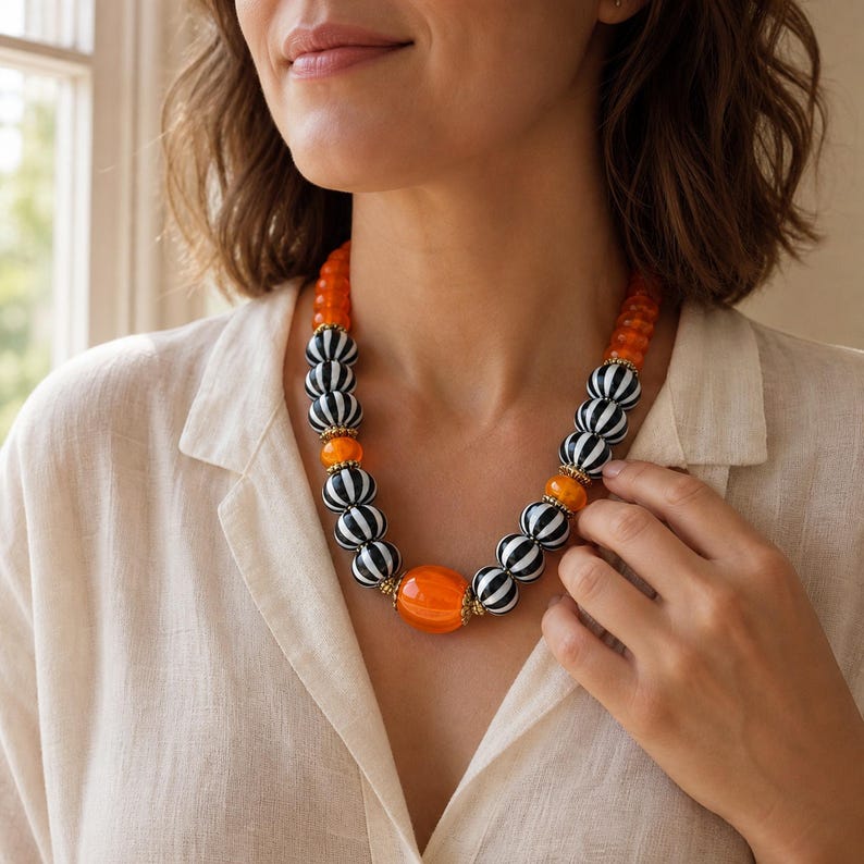 May include: A statement necklace featuring alternating round beads in vibrant orange and black-and-white striped patterns. The design includes small gold-toned spacers between the beads, with a larger, smooth orange bead centred at the bottom.