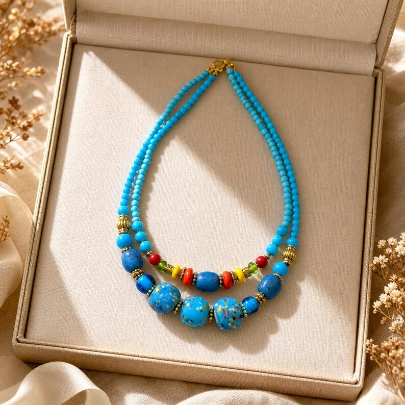 Puede incluir: Un collar de dos hileras con peque&ntilde;as cuentas de color turquesa, acentuado con cuentas azules moteadas m&aacute;s grandes, cuentas cil&iacute;ndricas de color rojo y amarillo vibrante, cuentas verdes peque&ntilde;as y espaciadores dorados dispuestos sim&eacute;tricamente.