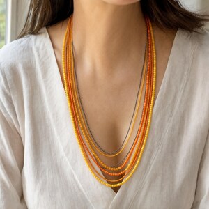 May include: A multi-strand necklace featuring several layers of small, round beads in shades of yellow, orange, and deep amber, interspersed with a single thin, dark metallic chain that hangs in a cascading, layered arrangement.