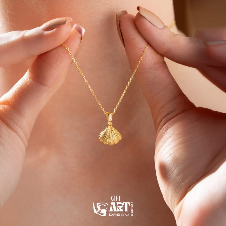 Op de afbeelding: Een goudkleurige ketting met een schelpenhanger. De hanger heeft een getextureerd oppervlak en een kleine, heldere edelsteen. De ketting wordt vastgehouden door de handen van een persoon. De tekst "GIFT ART DREAM" is ook zichtbaar.