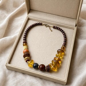 May include: A beaded necklace featuring a mix of dark brown wooden beads, translucent amber-coloured resin beads, and textured stone beads. The necklace includes small gold-toned metal spacers and a metal lobster claw clasp with an adjustable chain.