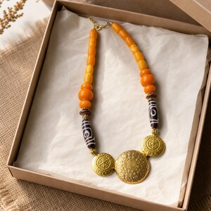 May include: A necklace featuring a strand of bright yellow and orange beads, two dark brown cylindrical beads with white spiral patterns, and three circular gold-toned metal pendants with intricate embossed geometric and circular designs.
