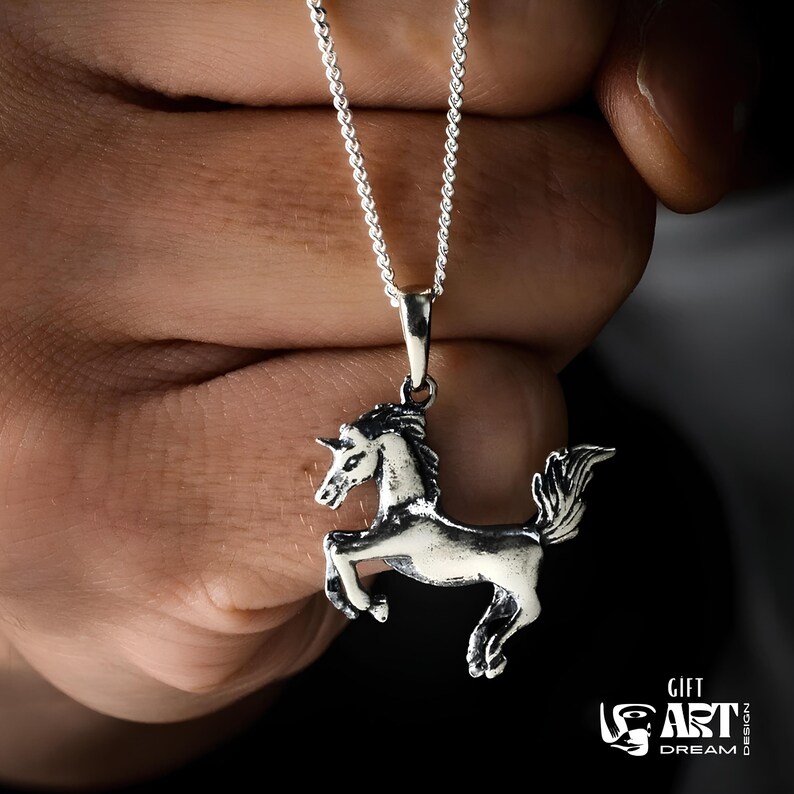 Puede incluir: Un collar con colgante de unicornio plateado. El colgante es una escultura tridimensional detallada de un unicornio con melena y cola fluidas. La cadena del collar es una delicada cadena de plata. El fondo es oscuro.
