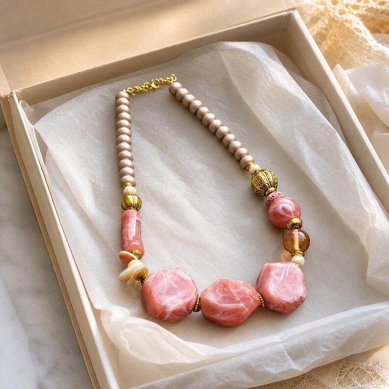 Pu&ograve; includere: Una collana composta da una fila di perline beige opache, arricchita da distanziatori in metallo dorato, sfere traslucide color ambra e grandi perle focali in resina rosa marmorizzata dalle forme irregolari e finitura liscia.