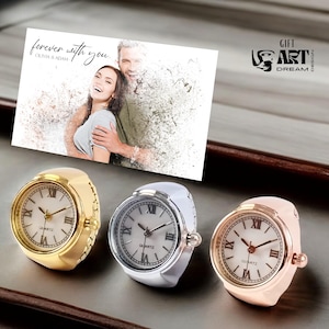 May include: Three ring watches with white faces and Roman numeral hour markers. One is gold-toned, one is silver-toned, and one is rose gold-toned. A card with the text "forever with you" and a couple's photo is in the background.