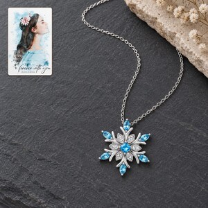 Winter Azure Snowflake Necklace styled against a dark luxury surface with the exact product finish clearly visible. A personalized digital greeting card is shown alongside.