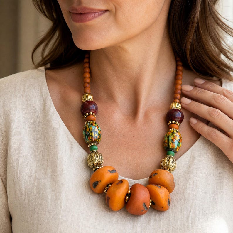 May include: A statement necklace featuring a mix of wooden, metallic, and glass beads. The design includes smooth orange amber-coloured stones, gold-toned filigree spheres, and multi-coloured speckled glass beads arranged on a string of brown beads.