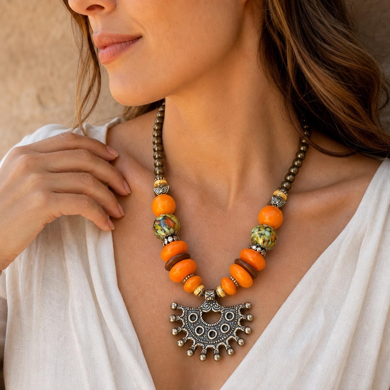 May include: A necklace featuring a series of round orange and brown beads interspersed with metallic accents and patterned green and orange beads, culminating in a large, semi-circular silver-toned pendant with intricate embossed circular details.