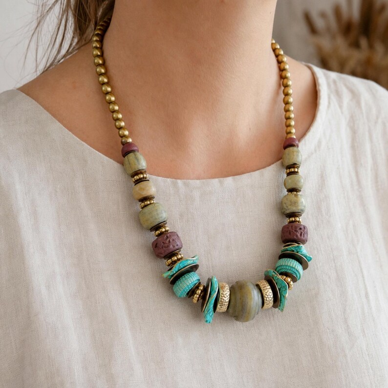 Puede incluir: Un collar de cuentas con una mezcla de esferas met&aacute;licas doradas, cuentas lisas de color verde salvia, cuentas granates texturizadas y acentos de color turquesa vibrante. El dise&ntilde;o incorpora varias formas y tama&ntilde;os de cuentas en un patr&oacute;n sim&eacute;trico.