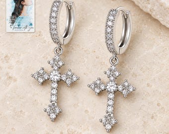 May include: A pair of silver-toned hoop earrings featuring a dangling cross pendant. Both the hoops and the cross pendants are encrusted with small, clear, sparkling stones. A small card in the corner displays an illustration of a person with the text "forever with you".