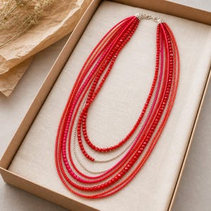 May include: A multi-strand necklace featuring various shades of red and pink beads. The necklace consists of several layered strands of small, round, and faceted beads in vibrant red, coral, and magenta tones, secured with a silver-toned metal clasp.