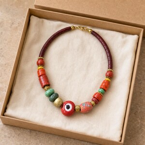 Red green and antique gold chunky necklace with central red evil eye bead worn with an ivory linen top in warm natural window light