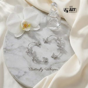 May include: A silver ear cuff with butterfly detailing, displayed on a marble surface alongside a white orchid and a small glass bottle. The ear cuff has multiple butterfly designs and the text "Butterfly Whisper".