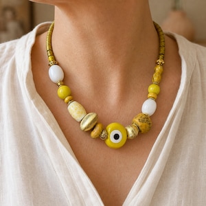 May include: A beaded necklace featuring a central yellow evil eye charm, interspersed with white, yellow, and textured gold-toned beads. The design includes metallic spacers and a series of small, cylindrical brass-colored beads along the neckline.