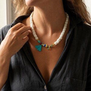 Puede incluir: Un collar de cuentas con cuentas blancas de forma cuadrada, acentuado con una borla turquesa, una cuenta de ojo turco multicolor, peque&ntilde;os espaciadores dorados y un peque&ntilde;o dije colgante, creando una apariencia texturizada y detallada.