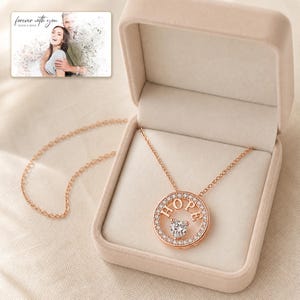 Puede incluir: Un collar con colgante circular en tono oro rosa que presenta la palabra HOPE en letras may&uacute;sculas y una piedra central en forma de coraz&oacute;n, rodeada de peque&ntilde;os cristales claros. El collar se muestra en una caja beige con una foto de una pareja.