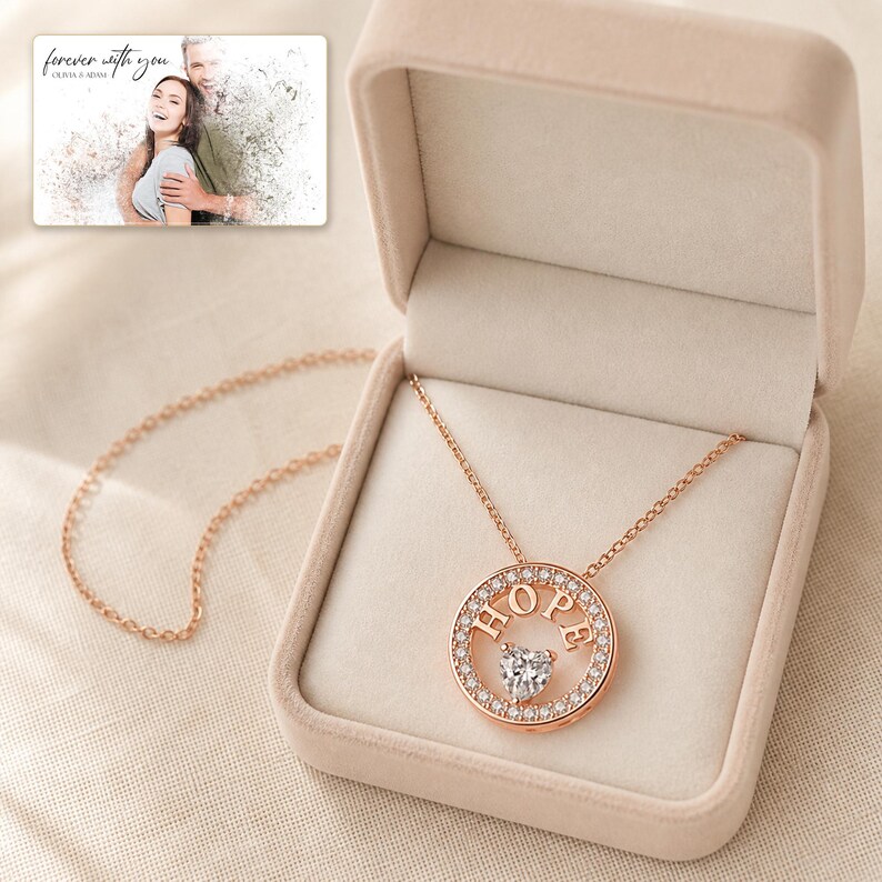 Puede incluir: Un collar con colgante circular en tono oro rosa que presenta la palabra HOPE en letras may&uacute;sculas y una piedra central en forma de coraz&oacute;n, rodeada de peque&ntilde;os cristales claros. El collar se muestra en una caja beige con una foto de una pareja.