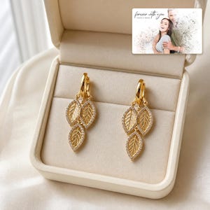 May include: A pair of gold-toned hoop earrings featuring three dangling leaf-shaped charms encrusted with small clear stones. The earrings are displayed inside a cream-colored jewelry box next to a small card with the text forever with you.