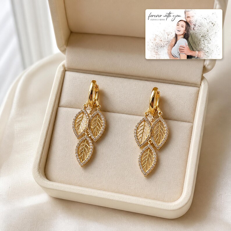 May include: A pair of gold-toned hoop earrings featuring three dangling leaf-shaped charms encrusted with small clear stones. The earrings are displayed inside a cream-colored jewelry box next to a small card with the text forever with you.