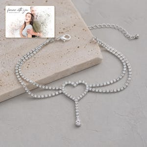 May include: A silver-toned anklet featuring two delicate chains adorned with clear rhinestones, a central open heart-shaped charm, and a single dangling rhinestone drop. The jewelry includes a lobster claw clasp and an adjustable extender chain.