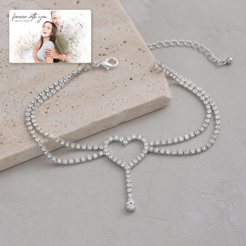 May include: A silver-toned anklet featuring two delicate chains adorned with clear rhinestones, a central open heart-shaped charm, and a single dangling rhinestone drop. The jewelry includes a lobster claw clasp and an adjustable extender chain.