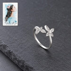 Double butterfly ring with clear accents displayed against charcoal slate. A personalized digital greeting card is shown alongside.