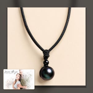 Black cord necklace with a dark round pendant, dark-bordered product view. A personalized greeting-card example appears in the corner.