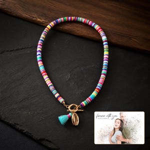 May include: A necklace made of multicolored flat circular beads featuring a gold-toned toggle clasp, a small light blue tassel, and a gold-toned shell charm. A rectangular card is included with the text "forever with you OLIVA & ADAM".