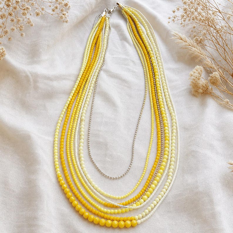 May include: A multi-strand necklace featuring various shades of yellow and cream beads. The design includes several layers of small, round beads in bright yellow, pale yellow, and off-white, accented by a single thin silver-toned metal chain.