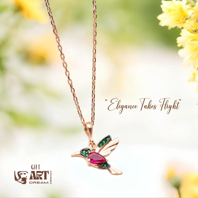 May include: A rose gold hummingbird pendant necklace with a ruby-colored gemstone and green accents. The necklace chain is delicate. The image includes the text "Elegance Takes Flight" and the logo "Gift Art Dream Design".
