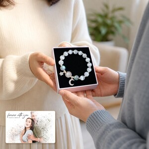 Puede incluir: Una pulsera de cuentas esf&eacute;ricas blancas y azuladas, con un dije de estrella y otro de media luna en tono plateado. La pulsera se presenta en una caja cuadrada. Una tarjeta rectangular muestra la foto de una pareja con el texto "forever with you" y "OLIVIA & ADAM".