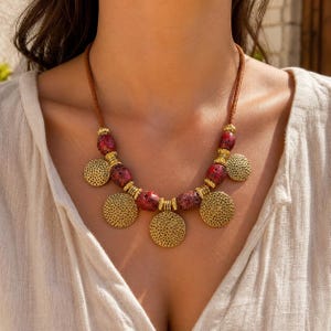 May include: A necklace featuring a brown cord, interspersed with textured red beads and gold-toned metal spacers. Five circular gold-toned pendants with a perforated, honeycomb-like pattern hang from the center, creating a layered, symmetrical design.