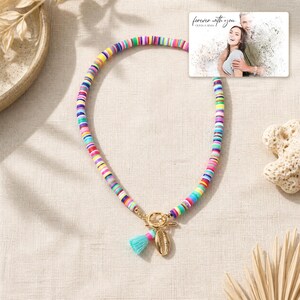 May include: A necklace made of small, multicolored flat circular beads featuring a gold-toned toggle clasp, a small turquoise tassel, and a gold-toned cowrie shell charm. A rectangular card displays a photo of a couple with the text "forever with you".