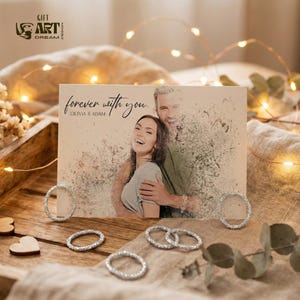 May include: A greeting card with a photo of a couple, the words "forever with you" and the names Olivia & Adam. Several silver rings and wooden hearts are arranged around the card on a wooden tray. Fairy lights are in the background.