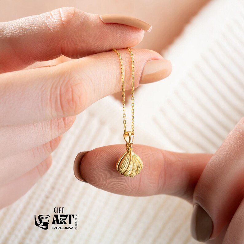 Op de afbeelding: Een goudkleurige ketting met een schelpenhanger. De hanger heeft een gedetailleerd, gestructureerd ontwerp. De ketting wordt door een persoon omhoog gehouden. De afbeelding bevat ook de tekst "GIFT ART DREAM DESIGN".