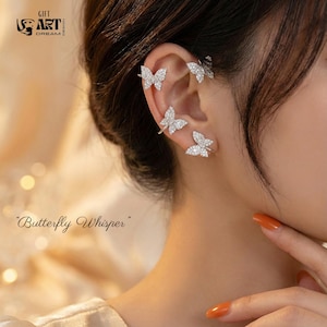 Puede incluir: Puños de oreja de mariposa plateados con detalles brillantes. El texto "Butterfly Whisper" y el logotipo "GIFT ART DREAM" son visibles. Un accesorio delicado y elegante para la oreja.