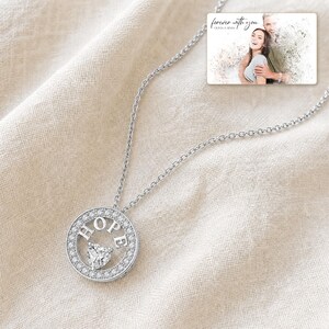 Puede incluir: Un collar de tono plateado con un colgante circular que presenta la palabra HOPE en letras may&uacute;sculas y un peque&ntilde;o cristal en forma de coraz&oacute;n en el centro. El colgante est&aacute; rodeado de peque&ntilde;as piedras claras. Una tarjeta rectangular muestra a una pareja y el texto forever with you.