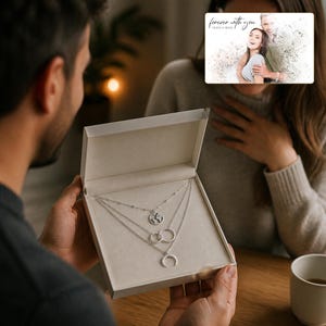May include: A silver-toned layered necklace set presented in a light-coloured gift box. The set features three distinct chains: one with a small globe pendant, one with two interlocking rings, and one with a crescent moon charm. The box is open.