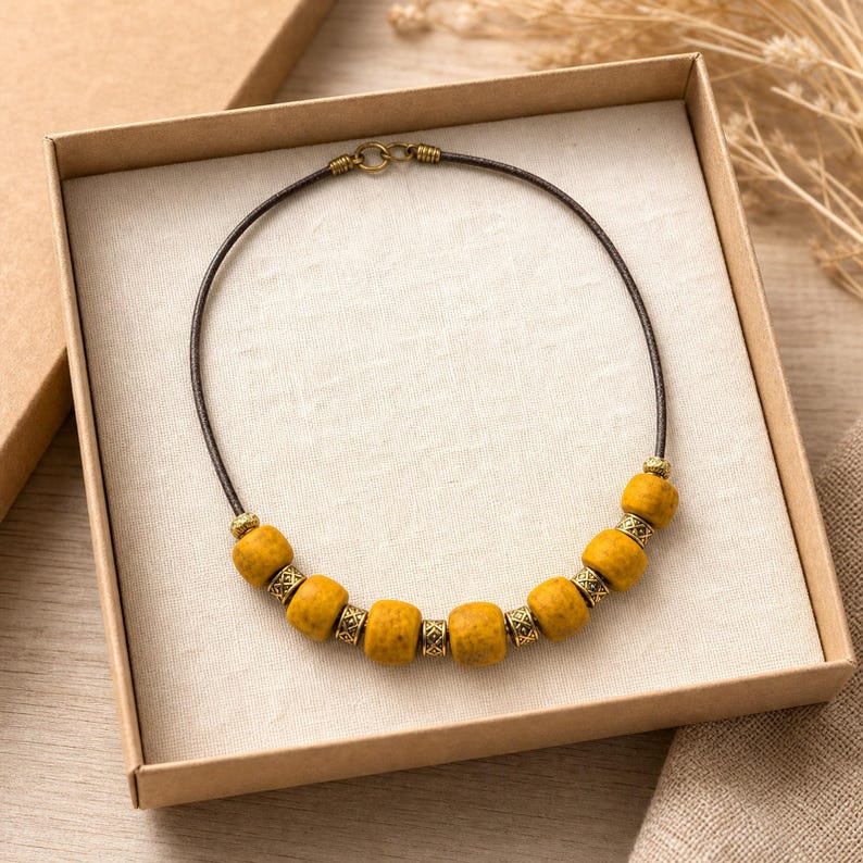 May include: A necklace featuring a dark brown cord with a series of mustard yellow, textured, rounded beads alternating with smaller, intricate, antique brass-toned metal spacers. The necklace includes a brass-toned metal clasp at the top center.