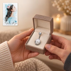 Two adults exchange an open taupe styling box with one white-ball skeleton-claw barbell visible inside. A personalized digital greeting card is shown alongside.