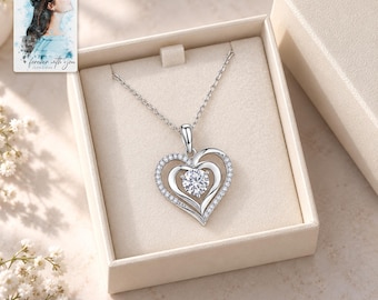 May include: A silver-toned heart-shaped pendant necklace featuring a central round clear stone surrounded by a pavé-set border. The necklace is presented in a beige box alongside a small card with an illustration of a person and the text "forever with you".