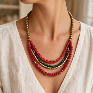 May include: A multi-strand necklace featuring layers of red, dark green, and metallic gold beads. The design includes smooth round gold beads at the top, transitioning into faceted red and green beads, interspersed with small square gold-toned spacers.