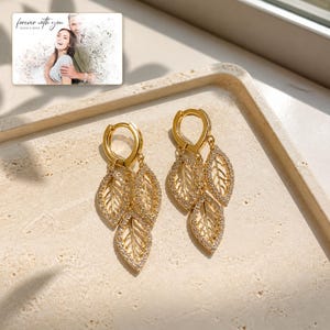 May include: A pair of gold-toned hoop earrings featuring three dangling leaf-shaped charms encrusted with small clear stones. The earrings are displayed alongside a small rectangular card with the text "forever with you" and "OLIVIA & ADAM".