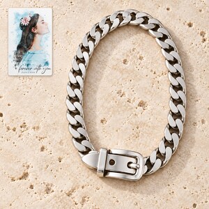 Polished silver tone curb-chain bracelet with belt-style buckle arranged vertically on ivory travertine, showing the complete chain and clasp. A personalized digital greeting card is shown alongside.