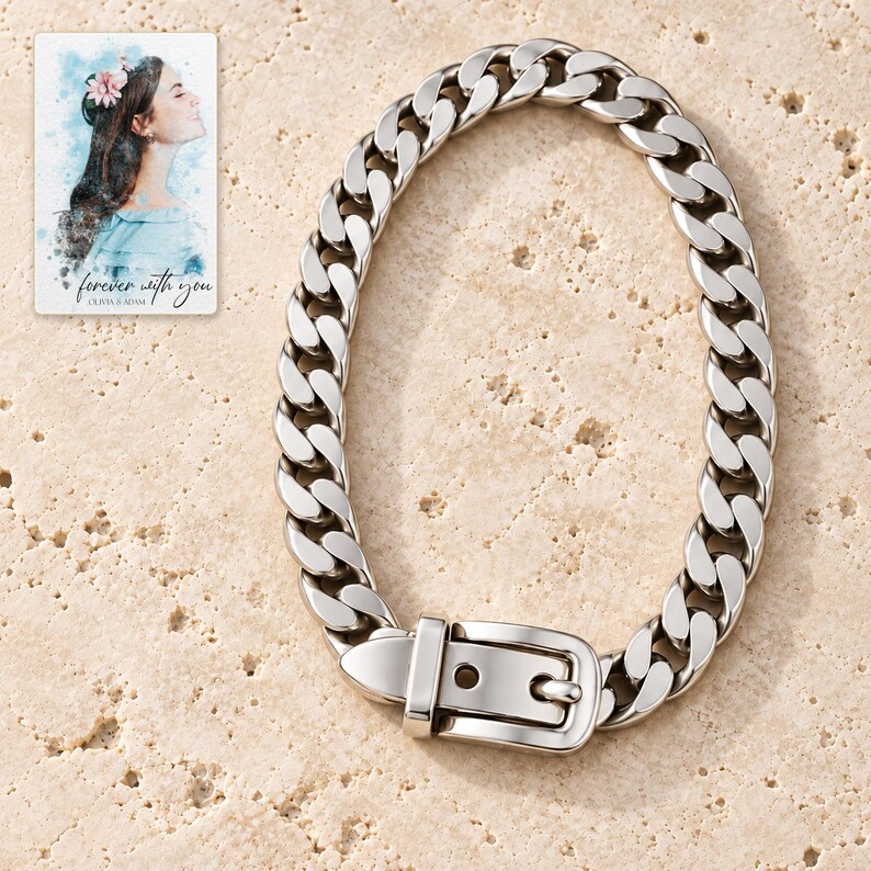 Polished silver tone curb-chain bracelet with belt-style buckle arranged vertically on ivory travertine, showing the complete chain and clasp. A personalized digital greeting card is shown alongside.