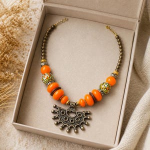 Pode incluir: Um colar com um pendente central de metal com acabamento antigo e design semicircular festonado. A corrente &eacute; composta por contas met&aacute;licas, contas lisas de cor laranja vibrante e contas salpicadas multicoloridas em tons de verde e laranja.