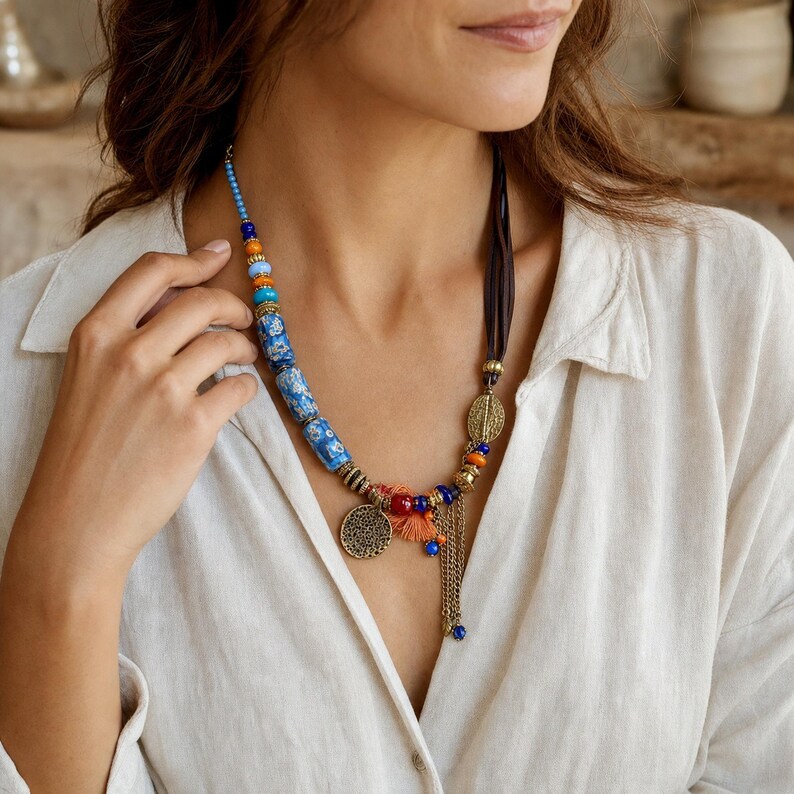 May include: A multi-strand necklace featuring blue and white patterned cylindrical beads, small orange and blue round beads, and metallic gold-toned accents. The piece includes a circular filigree charm, an orange tassel, and a dangling gold chain.