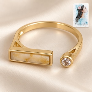 May include: A gold-toned open ring featuring a rectangular bar inlaid with a cream and tan marbled stone on one end and a single round clear gemstone on the other. A small rectangular card with a watercolor illustration of a person and the text "forever with you" is visible in the corner.